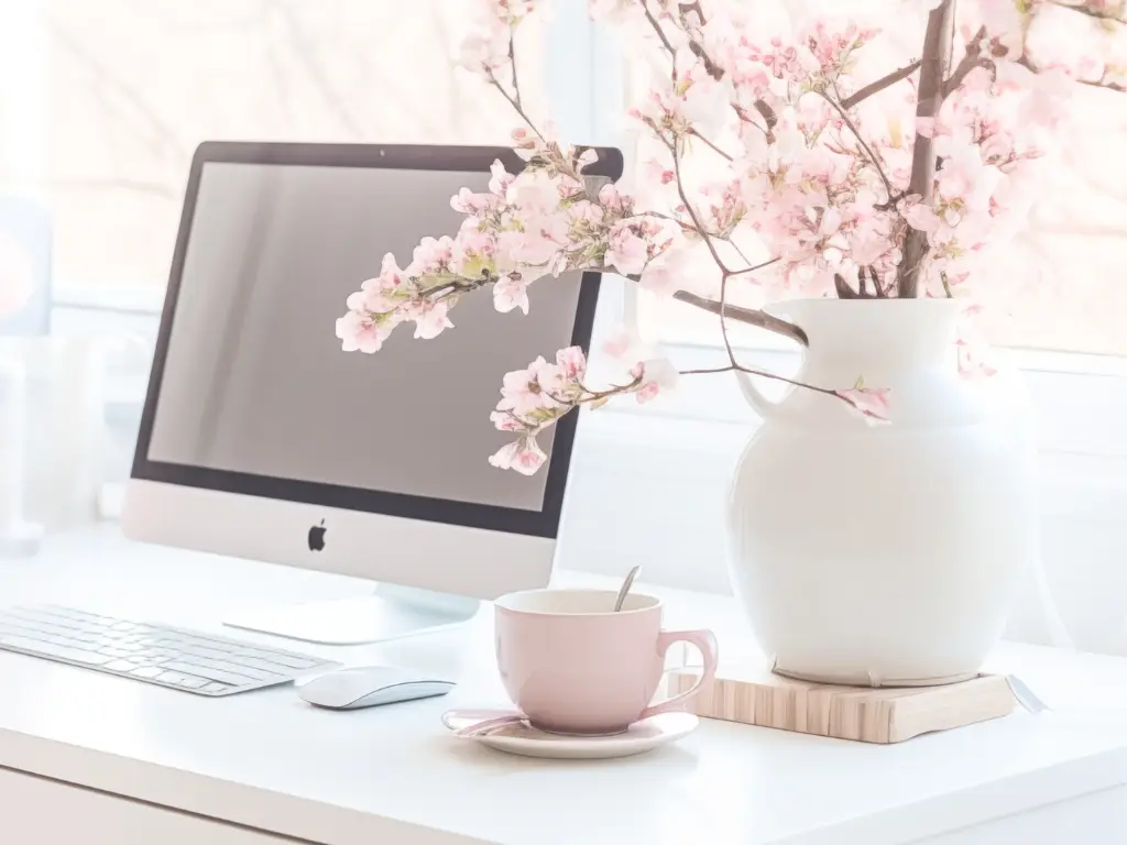 A Computer Desk with Flowers, coffee, monitor, keyboard and mouse.